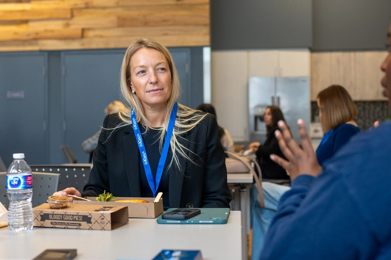 A woman with a blue lanyard sits at a table with food, listening to someone speaking across from her.