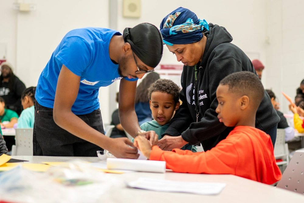 Two adults help two young boys with a craft activity at a table in a brightly lit classroom.