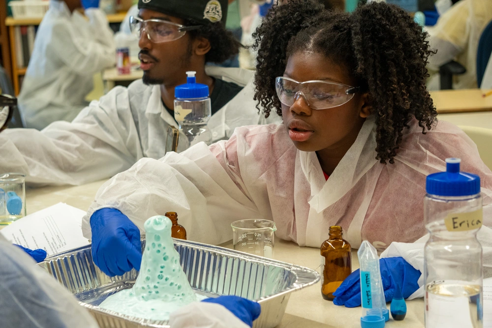 Two students wearing lab coats and goggles watch a foaming science experiment in a classroom setting.