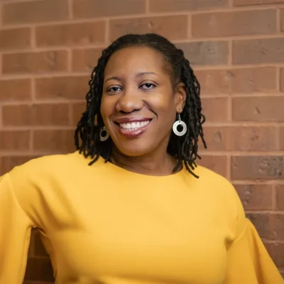 Smiling woman with medium-length locs in a yellow top standing in front of a brick wall.