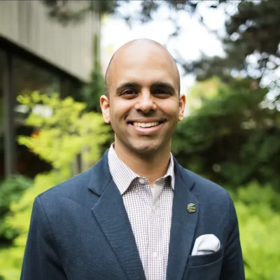 A smiling man in a blue blazer stands outdoors with greenery in the background.