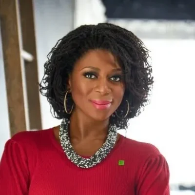 Woman with curly hair in a red top and statement necklace, smiling at the camera against a bright background.