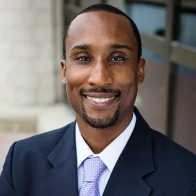 Man in a dark suit and light purple tie smiling outdoors in front of a building with glass windows.