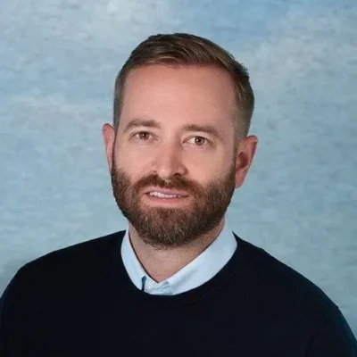 Man with short brown hair and beard wearing a navy sweater over a collared shirt, against a blue backdrop.