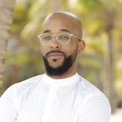 A man with glasses and a beard wearing a white shirt, standing outdoors with palm trees in the background.