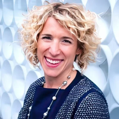 Smiling woman with short curly blonde hair, wearing a navy outfit and a beaded necklace, standing by a patterned wall.