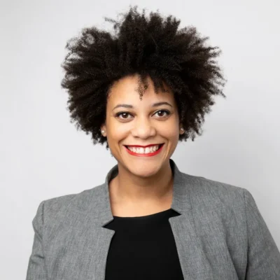 Smiling woman with curly hair in a gray blazer and black top against a plain white background.