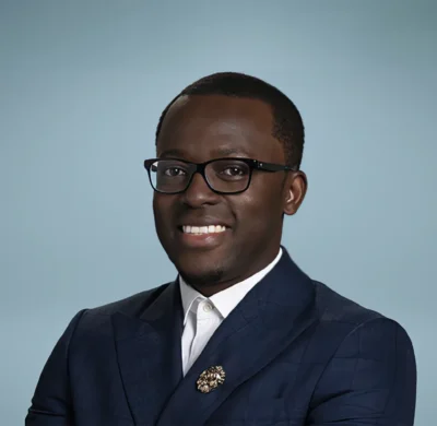 Smiling person in a dark suit and glasses against a plain light blue background.