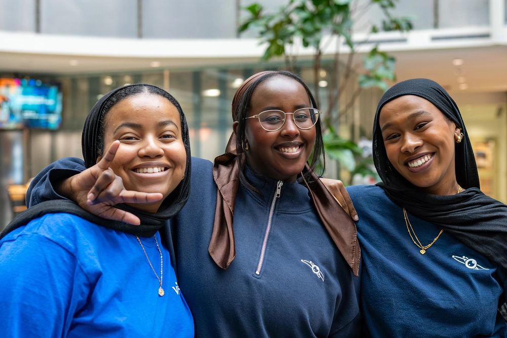 Three smiling women in hijabs pose together indoors, looking happy and friendly.