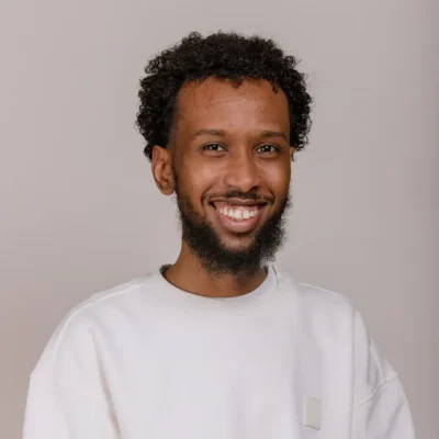 A smiling man with curly hair and a beard wearing a white sweatshirt against a plain background.