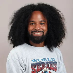 Smiling man with long, curly hair wearing a gray World Series sweatshirt against a plain background.
