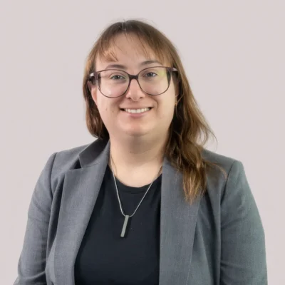 Person with long light brown hair and glasses, wearing a gray blazer and black top, smiling at the camera.
