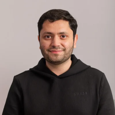 A man with short dark hair and a beard, wearing a black GUESS hoodie, smiles against a light gray background.