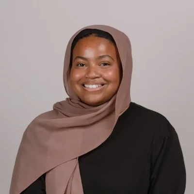 A woman wearing a taupe hijab and black top smiles at the camera against a plain background.