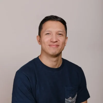 Man with short dark hair wearing a navy t-shirt, smiling in front of a plain light background.