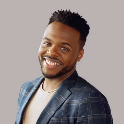 Smiling man with short curly hair in a plaid blazer and beige shirt, posing against a plain background.