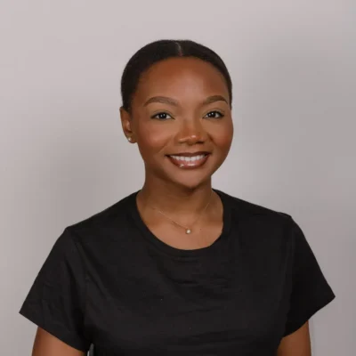 Smiling woman with short hair wearing a black shirt and necklace, posing in front of a plain background.