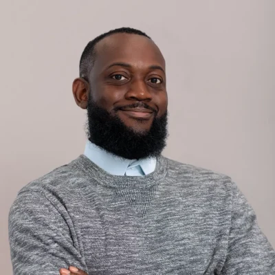 Man with a beard wearing a gray sweater and light blue shirt, smiling with arms crossed against a plain background.