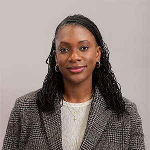 Woman with long braided hair wearing a plaid blazer, white top, and gold jewelry, posing against a gray background.