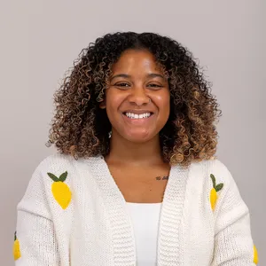 Smiling woman with curly hair wearing a white sweater with yellow lemon designs, against a plain background.