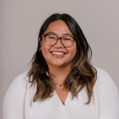 A woman with long dark hair, glasses, and a white sweater smiles in front of a plain light background.