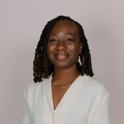A woman with twisted hair smiles, wearing a white blouse, gold necklace, and hoop earrings against a plain background.