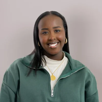 Woman with long dark hair, wearing a green zip-up sweater, white top, and gold jewelry, smiling at camera.