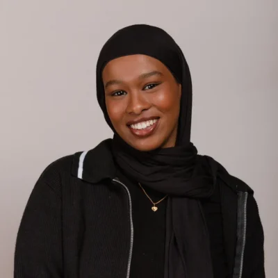 Smiling woman in a black hijab and jacket, wearing a gold necklace, against a plain background.