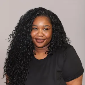 Woman with long curly black hair wearing a black shirt, smiling in front of a plain light background.
