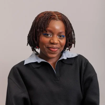 Smiling person with short locs, wearing a black sweater over a collared shirt, against a plain background.