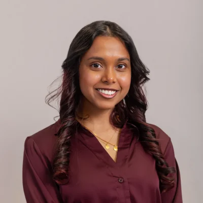 Smiling woman with long dark hair wearing a maroon blouse and gold necklaces, posed against a plain background.