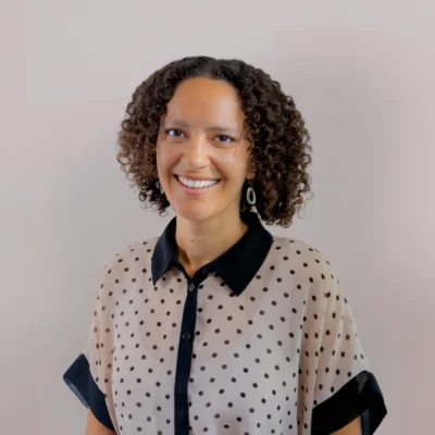 Smiling person with curly hair in a polka dot blouse standing against a plain light background.