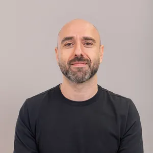 A man with a beard and shaved head wearing a black shirt, smiling slightly in front of a plain background.