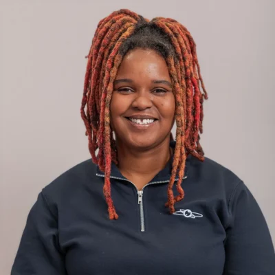 Smiling woman with red and orange locs wearing a navy zip-up top, standing against a plain background.