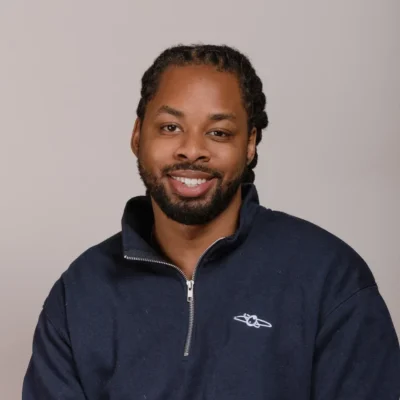 Man with braided hair and a beard, smiling, wearing a navy blue zip-up sweatshirt, sitting against a plain background.