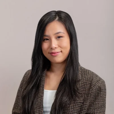 A woman with long dark hair in a brown checkered blazer smiles at the camera against a neutral background.