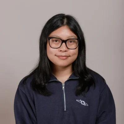 A person with long black hair and glasses, wearing a navy zip-up sweatshirt, smiles at the camera.