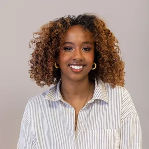 Smiling woman with curly hair wearing a striped shirt and gold hoop earrings against a plain background.