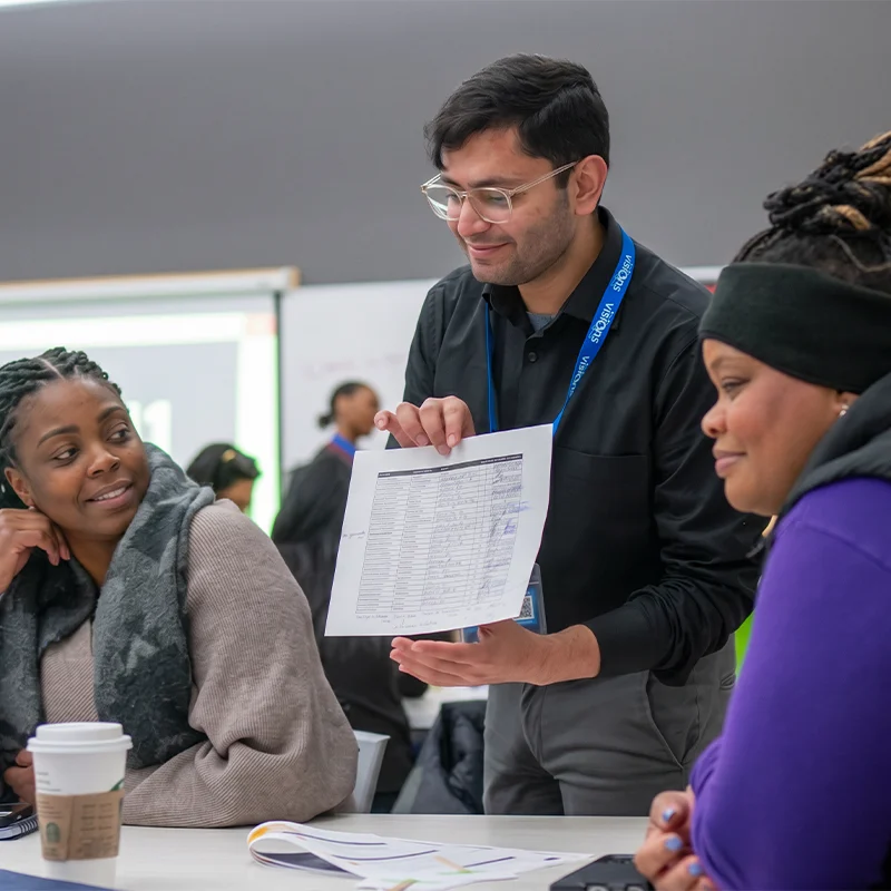 A man shows a printed document to two women seated at a table in a meeting or classroom setting.