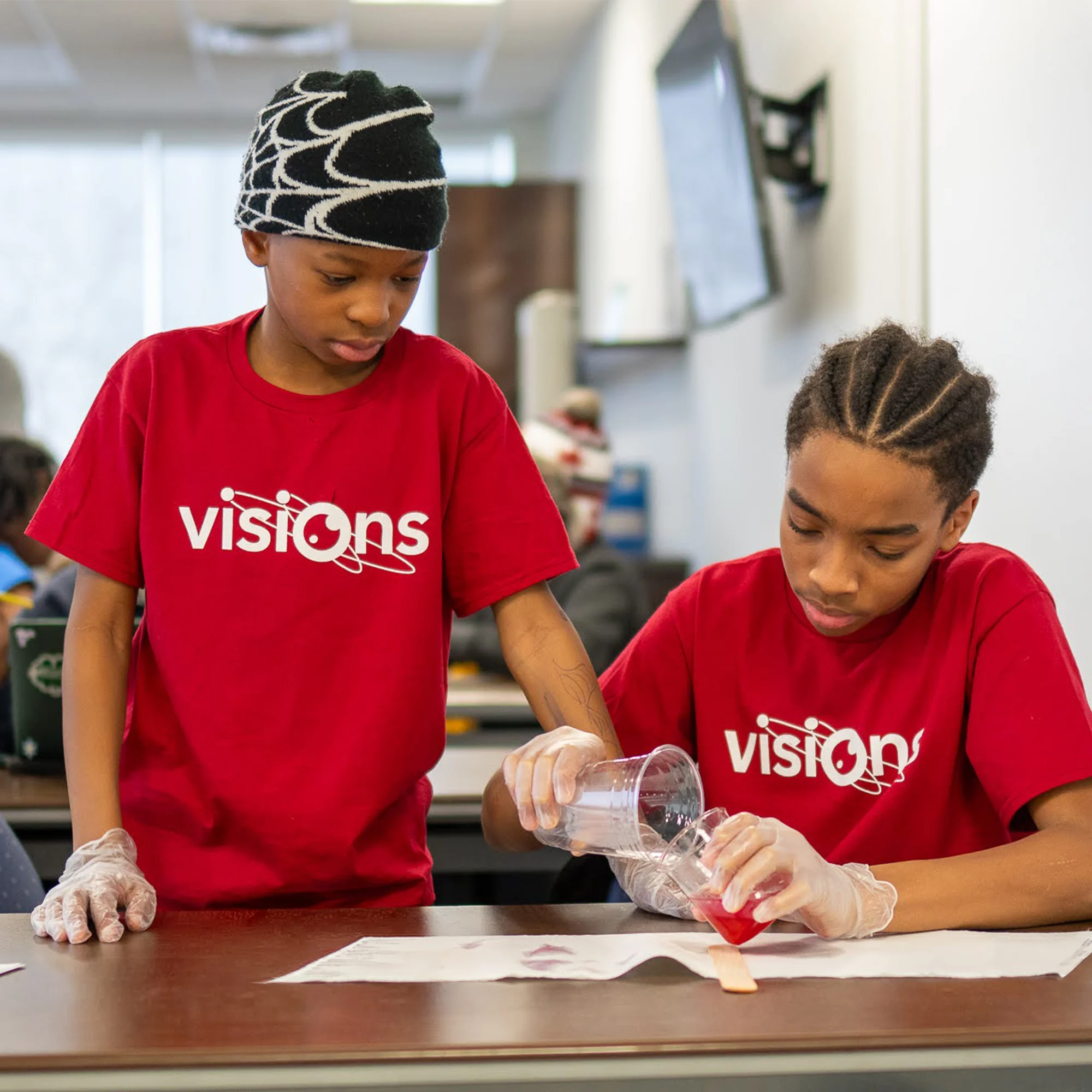 Two kids in red Visions shirts pour liquid into a cup during a classroom science activity.