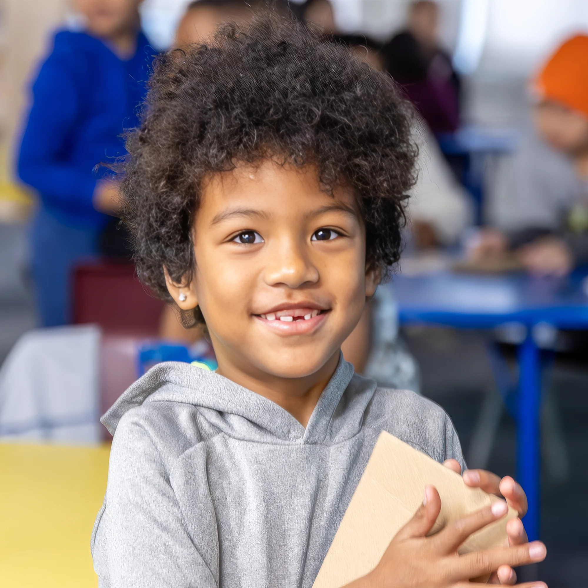 Smiling child holding a block in a classroom, with other children and desks blurred in the background.