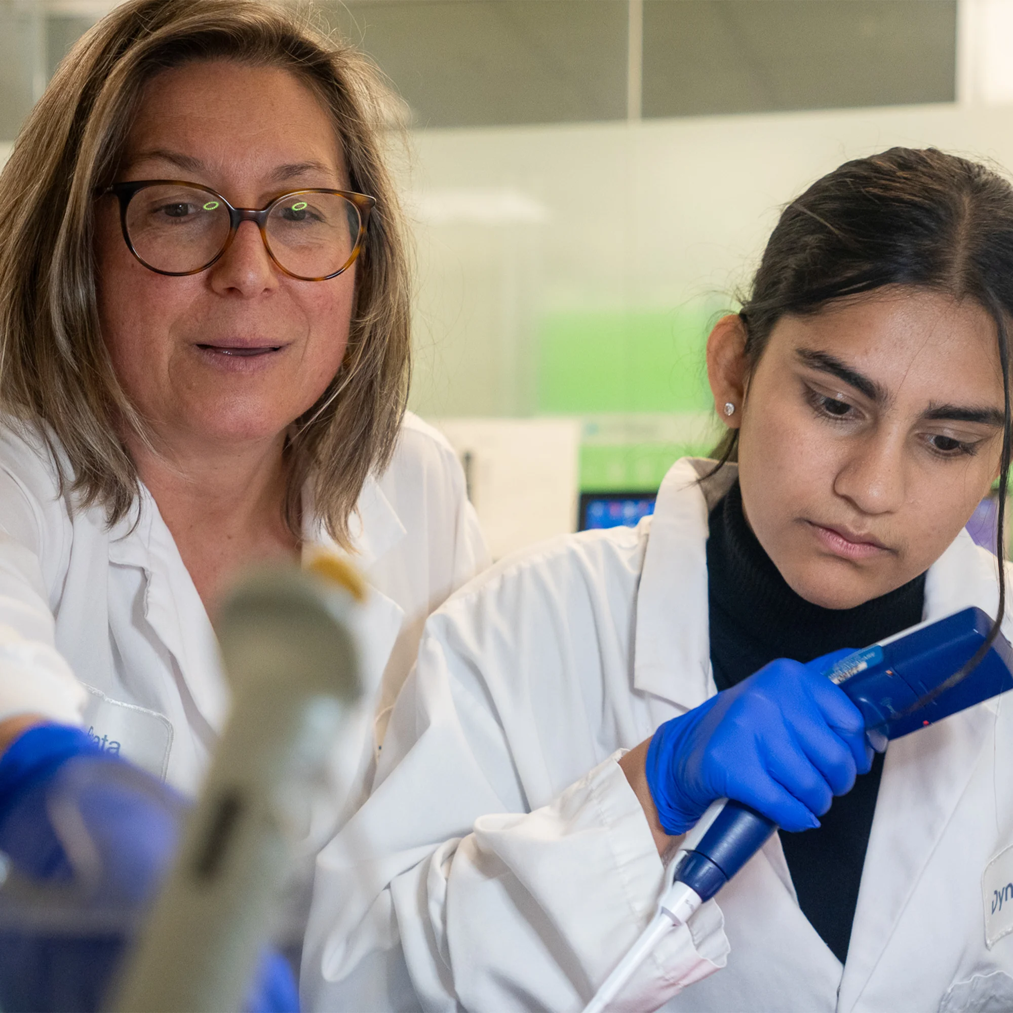 Two women in lab coats and gloves work with pipettes in a laboratory setting.