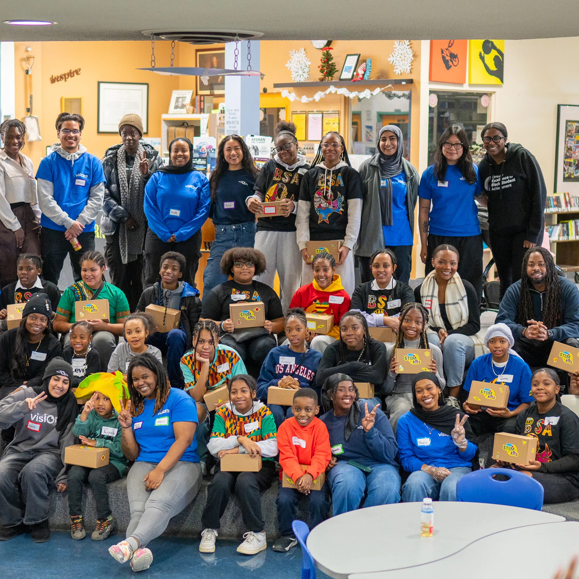 A large, diverse group of people and children pose together indoors, many holding decorated cardboard boxes and smiling.