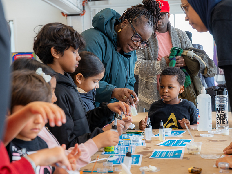 Adults and children gather around a table, engaging in hands-on science experiments with various materials.