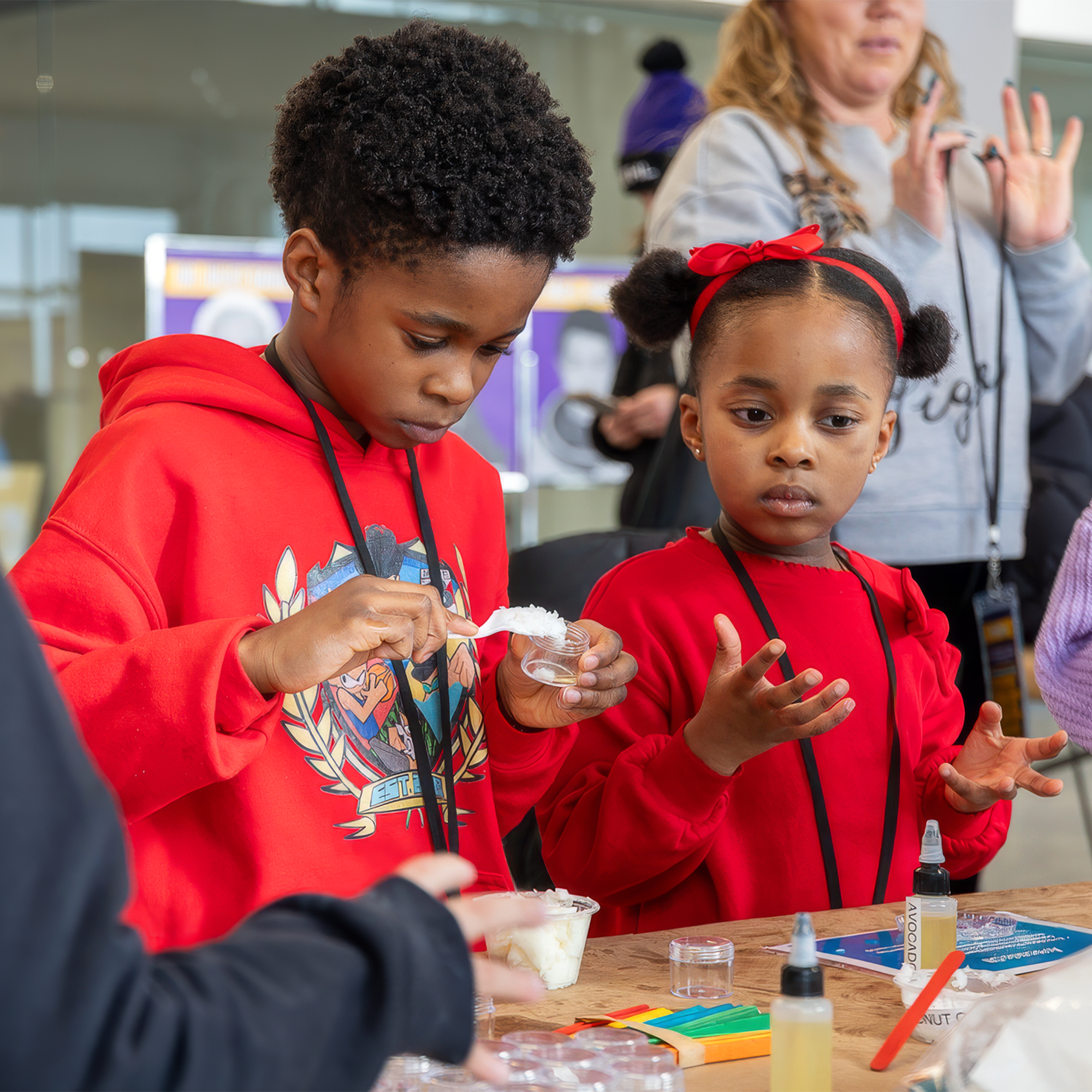 Two children do a science experiment at a table, surrounded by supplies, with adults watching in the background.