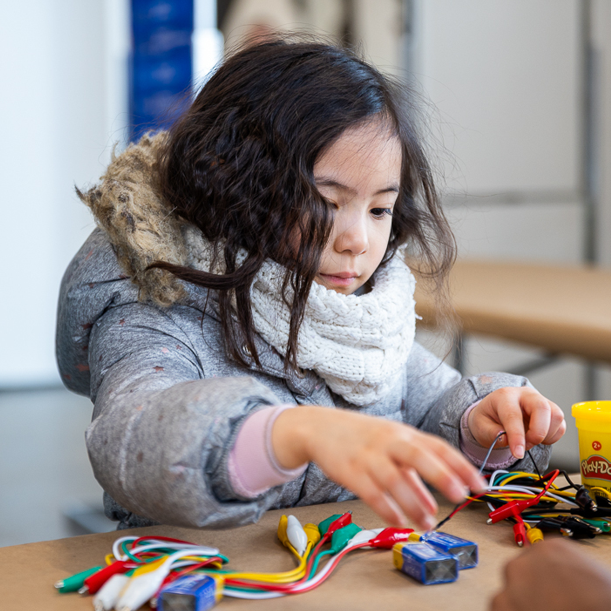 Young girl in a coat and scarf working with wires and batteries on a table, focused on a STEM project.
