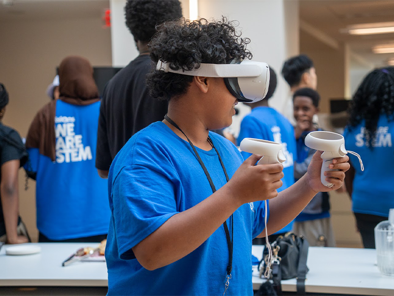 A student in a blue shirt uses a VR headset and controllers in a classroom with others in the background.