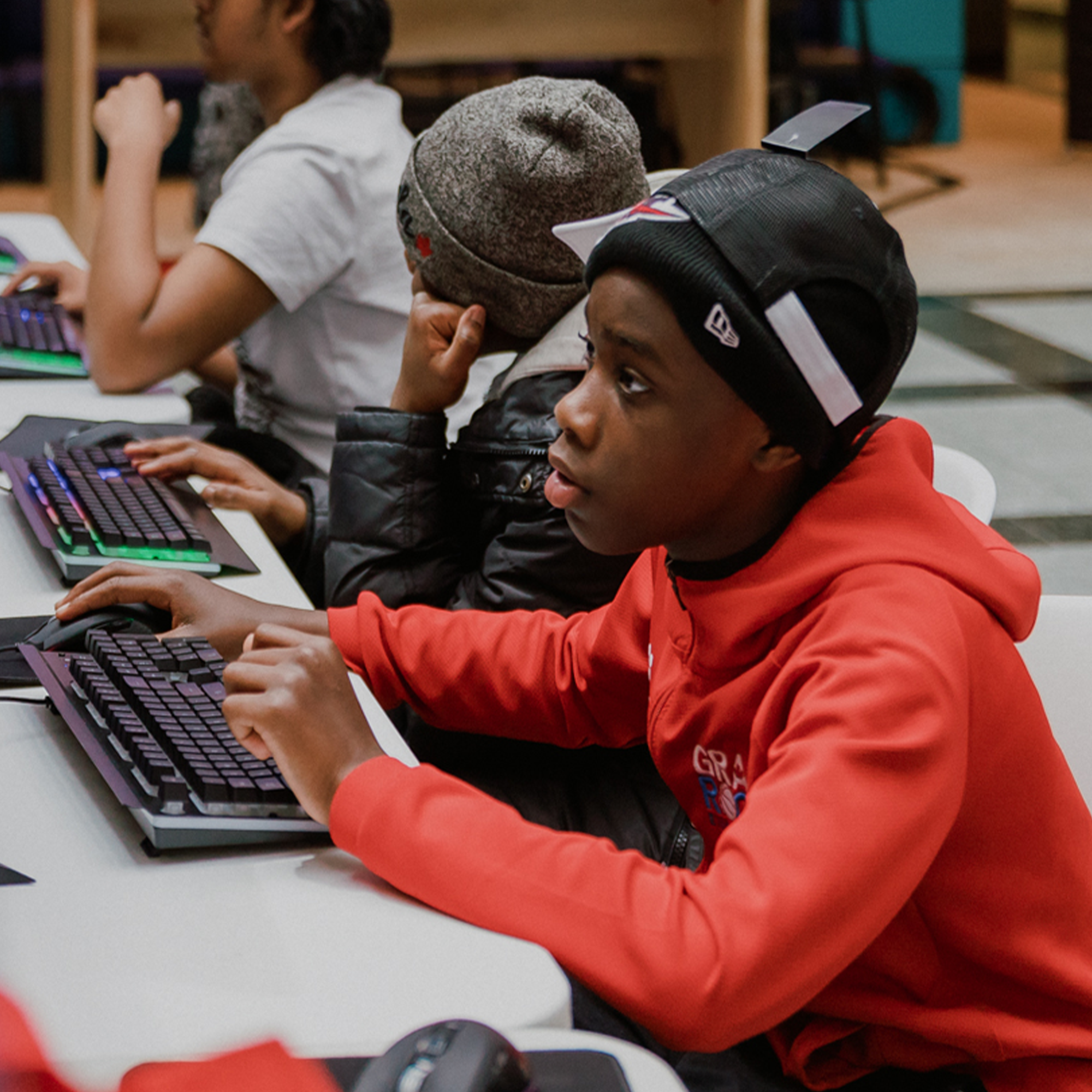 A boy in a red hoodie uses a computer keyboard, focused, with others beside him at a desk.