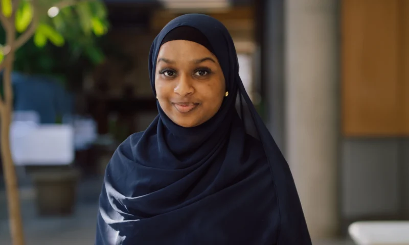 A woman in a navy hijab smiles softly while standing indoors with blurred background.