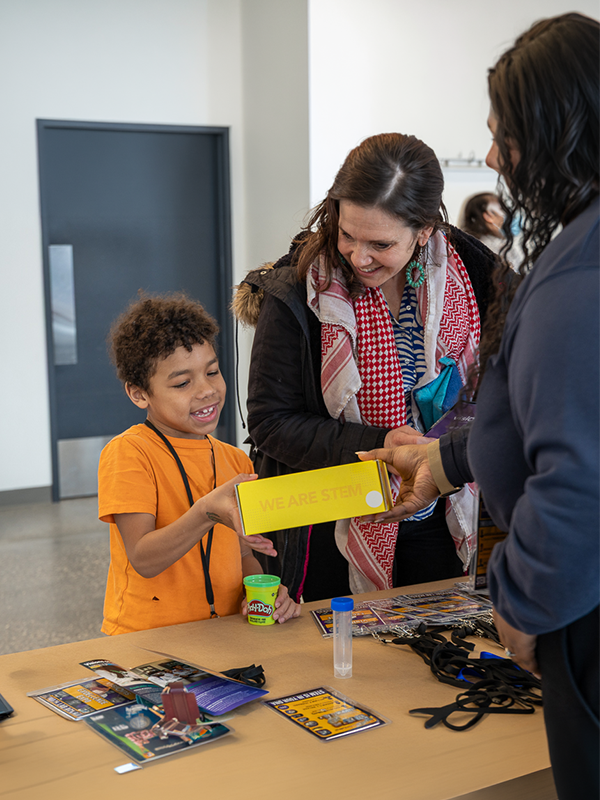 A smiling child receives a yellow We Are STEM box from an adult at a table with science materials.